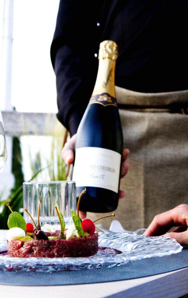 Cameriere in divisa che presenta una bottiglia di Champagne per l'abbinamento con una tartare gourmet, durante il servizio sulla terrazza del ristorante La Perla del Pescatore a Desenzano del Garda.