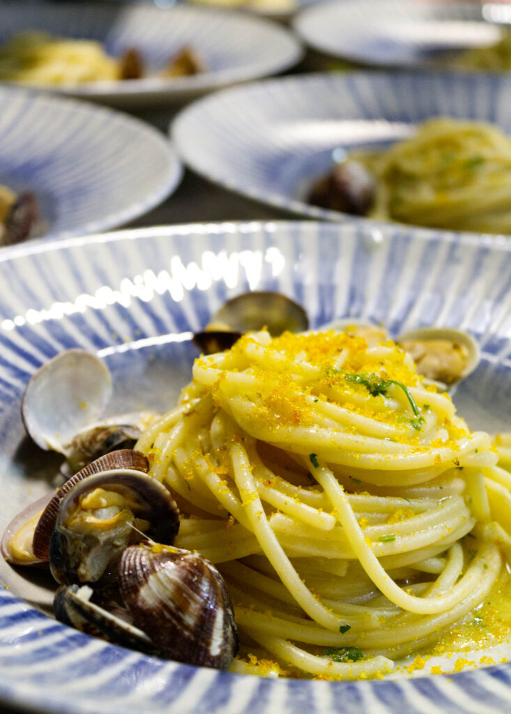 Primo piatto di spaghetti alle vongole veraci con prezzemolo e bottarga grattugiata, servito nei piatti di design del ristorante La Perla del Pescatore a Desenzano del Garda.