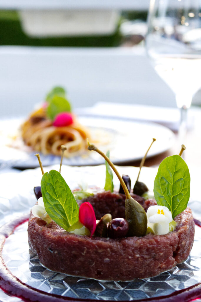 Inquadratura verticale di una tartare di cavallo gourmet con frutti di cappero e fiori eduli, con un primo piatto di pasta e un calice di vino sfocati sullo sfondo del ristorante La Perla del Pescatore a Desenzano del Garda.