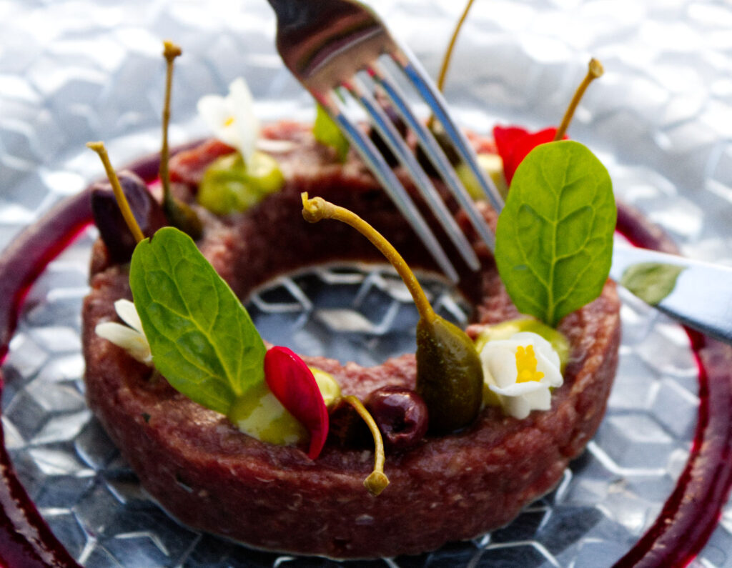 Antipasto gourmet di tartare di cavallo a forma di anello con frutti di cappero, olive, emulsione artigianale e fiori eduli, servito su piatto di vetro del ristorante La Perla del Pescatore a Desenzano del Garda.